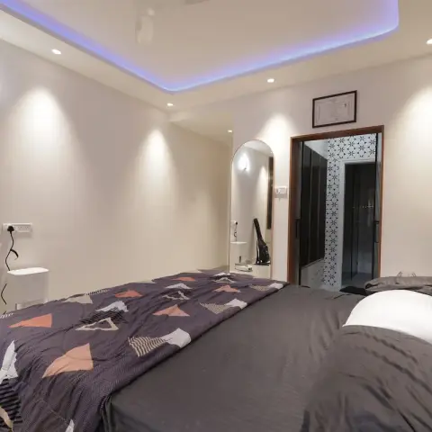 A grey-toned bedroom with a blue LED cove ceiling and a full-wall upholstered headboard, opening into an en-suite bath through a patterned glass door.