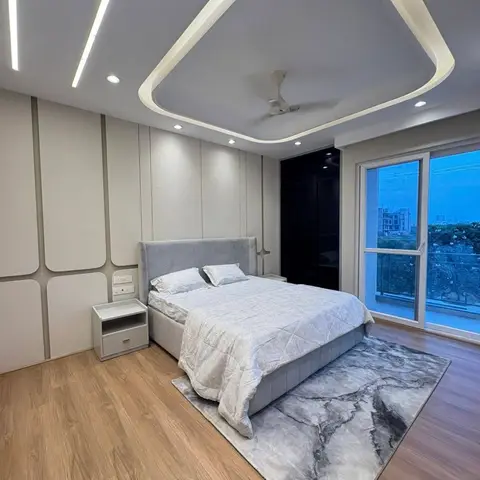 A grey tufted bed sits beneath a triangular LED-lit cove ceiling in a wood-floored bedroom with a glossy black wardrobe and a glass sliding door opening to a