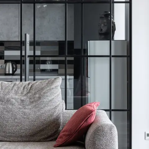 A grey tweed sofa with a red accent cushion in front of a black-framed glass partition opening to a mosaic-tiled kitchen.