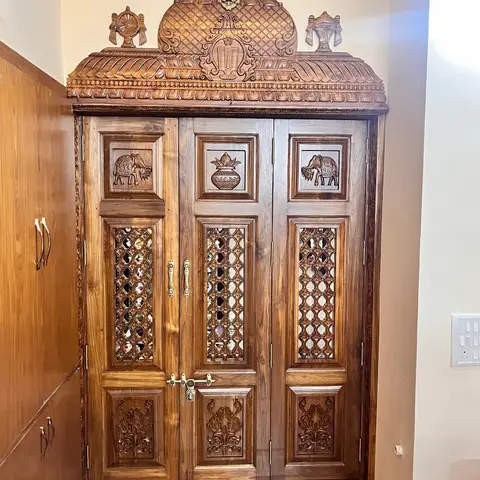 A hand-carved solid-wood pooja unit door with a temple-style pediment, elephant and kalash motifs, and mirror-inlaid jaali panels.