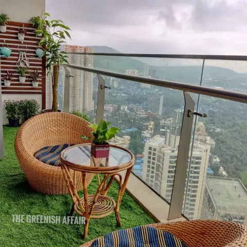 A high-rise balcony with rattan bistro seating on artificial turf, a vertical wall planter, and glass railings overlooking the city skyline.