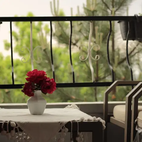 A home balcony with a wrought-iron scrollwork railing, hanging planters, a small table dressed with a tasseled cloth and a vase of red roses