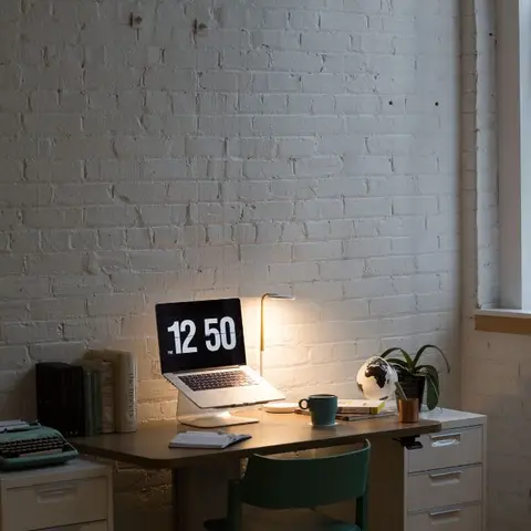 A home-office corner with a wooden desk on white filing pedestals against a painted white exposed-brick wall, lit by an arc lamp.