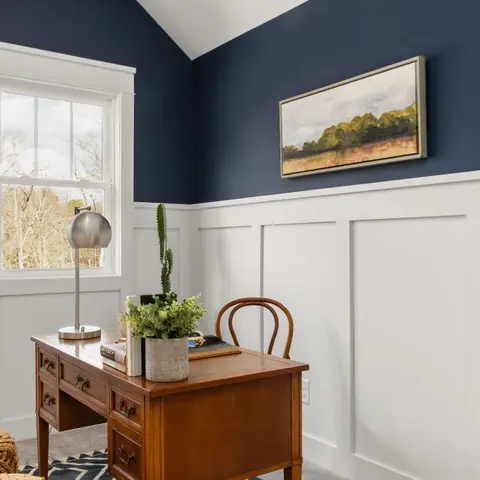A home study corner with navy upper walls above white wainscot panelling, a vintage wood writing desk with a bentwood chair by the window