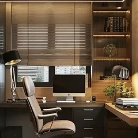 A home study with a built-in wood desk and open shelving unit, a black task lamp, and woven roman blinds over the window.