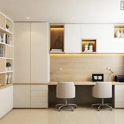 A home study with a built-in two-person desk under warm-lit display niches, a tall wardrobe cabinet, and an open bookshelf styled with books and plants.