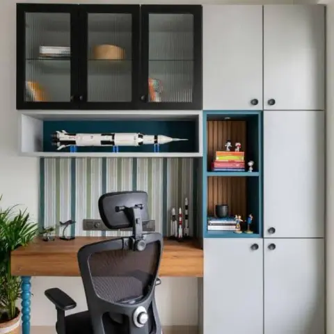 A kid's study nook with grey cabinetry, a blue-accented display niche styled with a rocket model and books, and striped wallpaper behind a wood desk.