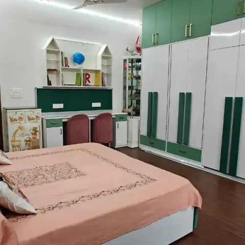 A kids bedroom has a house-shaped bookshelf and green study desk beside a green-and-white wardrobe, with a peach bedspread in the foreground.