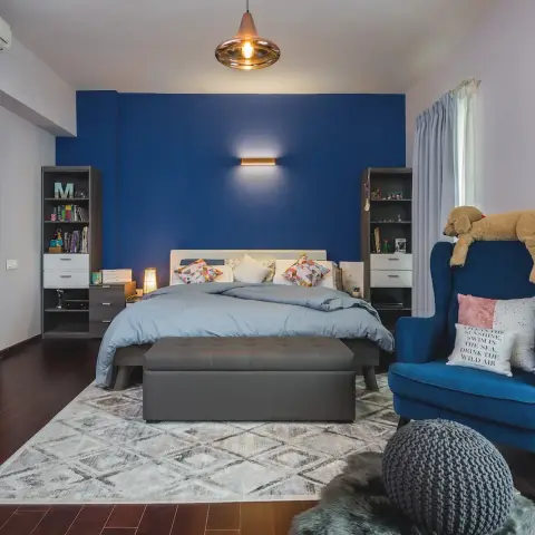 A kids' bedroom with a bold cobalt-blue accent wall, a navy wingback armchair styled with a plush toy, and a monogrammed bookshelf beside the bed.