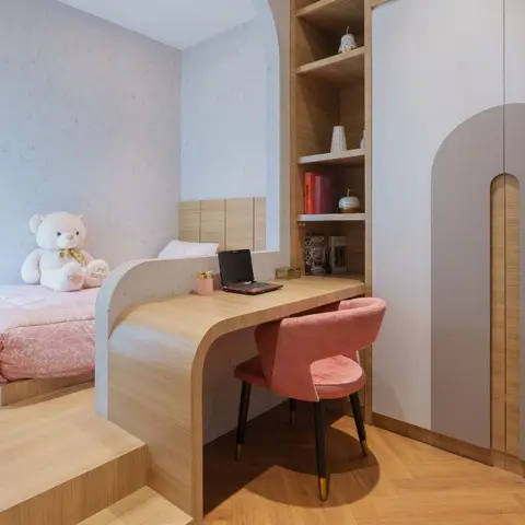 A kids' bedroom with a curved built-in study nook beside a platform bed, an arched wardrobe door, and a pink accent chair on herringbone wood flooring.