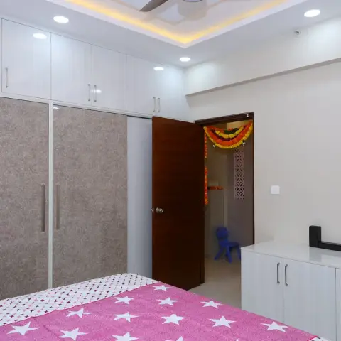 A kids' bedroom with a pink star-print bedspread, a floor-to-ceiling textured wardrobe, and a cove-lit ceiling, with a doorway open to an adjoining room.