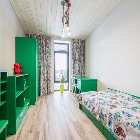 A kids' bedroom with bright green built-in wardrobe, shelving and desk, a single bed with car-print bedding, patterned curtains