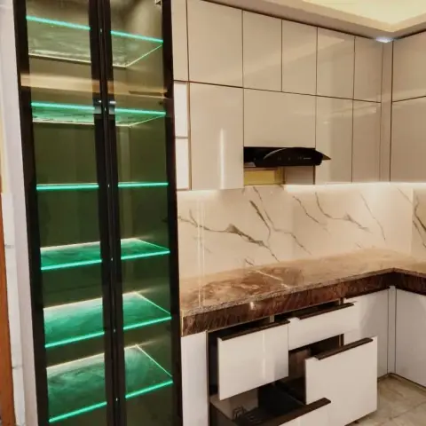A kitchen with a green LED-lit glass-front crockery display cabinet beside white gloss cabinetry and a brown-veined marble countertop and backsplash