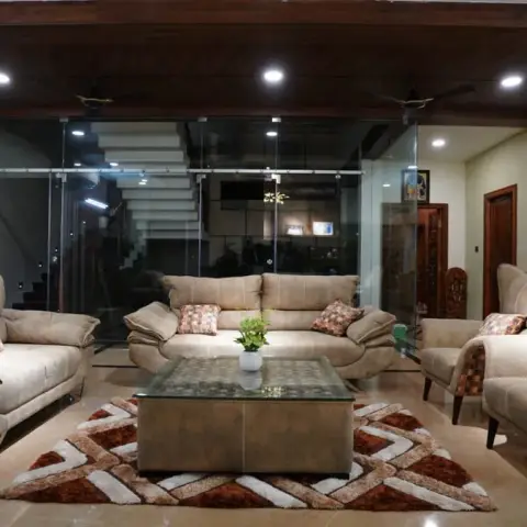 A large living room with a dark wood beam ceiling, beige sofas and wingback chairs around a glass coffee table on a chevron rug