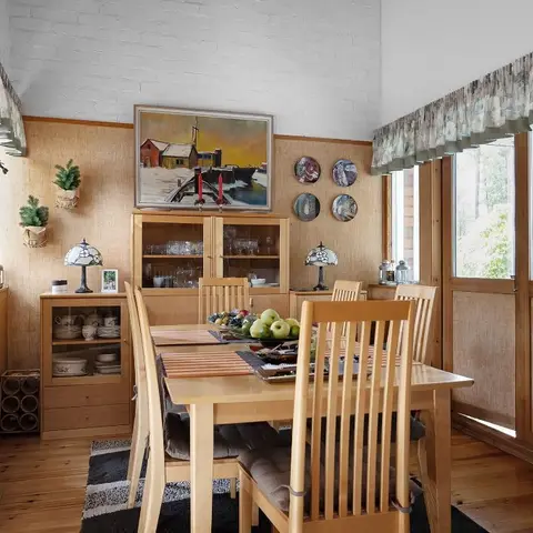 A light wood dining table and slat-back chairs beneath a framed harbour painting, next to a glass-front crockery cabinet displaying glassware and plates.