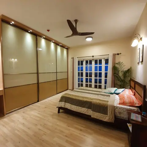 A lived-in bedroom with a full-wall backlit sliding wardrobe in cream and bronze glass panels, a wood bed and French doors opening to a balcony.