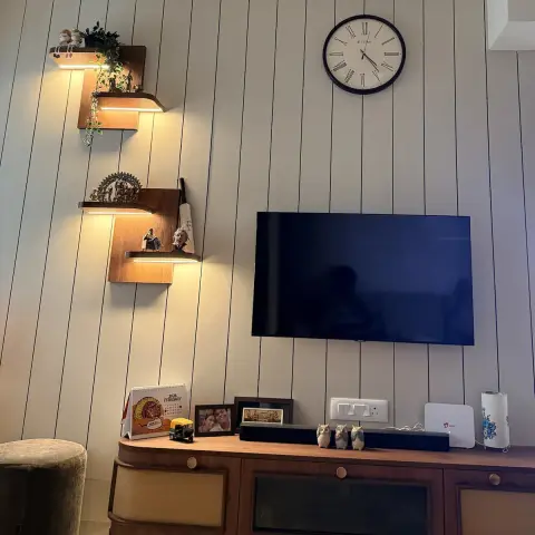 A lived-in TV wall of cream grooved panelling with a Roman-numeral clock, three stepped walnut display ledges lit by strip lights holding a Nataraja and family