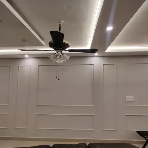 A living room accent wall with full-height panel moulding beneath a layered cove-lit false ceiling and an ornate ceiling fan with frosted glass shades.