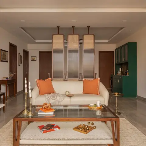 A living room anchored by a cream sofa with rust-orange cushions, a cane-and-reeded-glass room divider screen, and a forest green built-in bar cabinet.
