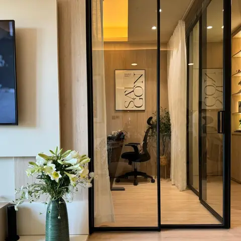 A living room console styled with a vase of white lilies beside a wall-mounted TV, with a glazed sliding-door study visible beyond a sheer curtain