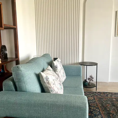 A living room corner with a sage-green sofa and embroidered floral cushions against a fluted wall panel, beside an arched wall niche and a vintage-pattern rug