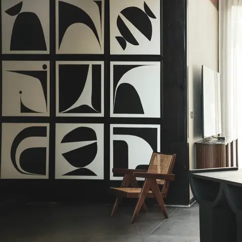 A living room corner with a nine-panel grid of black-and-white abstract wall art on a charcoal wall, a cane-and-wood armchair in Chandigarh-chair style