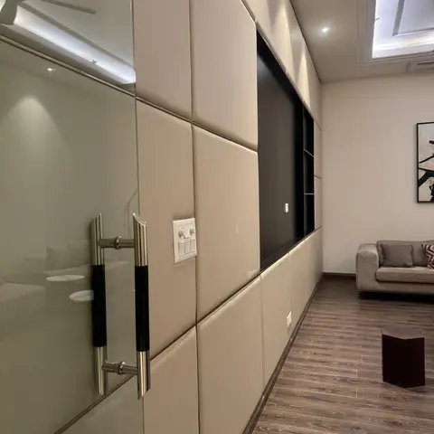 A living room feature wall of beige channel-tufted leather panelling frames a black gloss TV niche, seen past a glass pivot door with chrome handles.