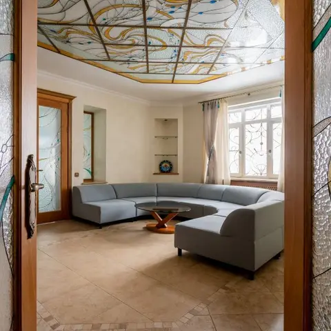 A living room framed by ornate art-nouveau stained-glass doors, with a matching stained-glass ceiling panel above a curved grey sectional sofa and a round wood