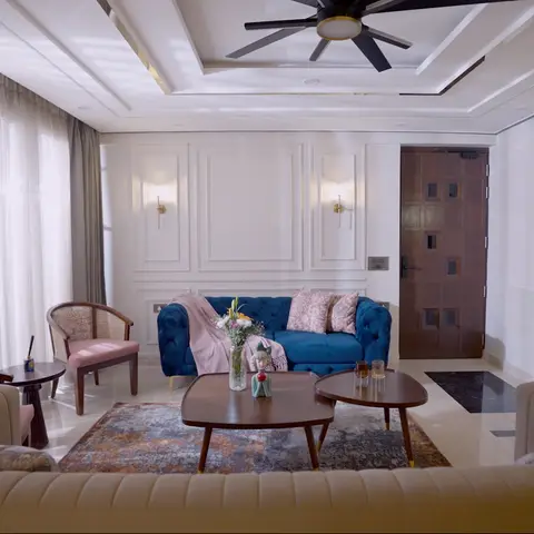 A living room mixing a tufted navy-blue velvet sofa, cane-backed armchairs, and twin wood coffee tables in front of a panelled wall and a carved wood door.