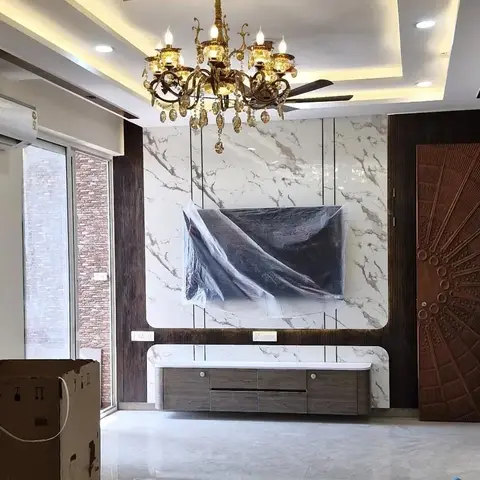 A living room TV wall clad in book-matched marble beneath a crystal chandelier, with a carved sunburst wooden door alongside