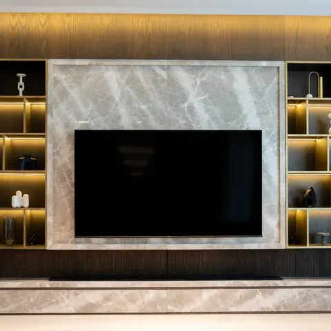 A living room TV wall in dark wood veneer with a book-matched marble inset panel, flanked by brass-edged backlit display shelving styled with ceramics