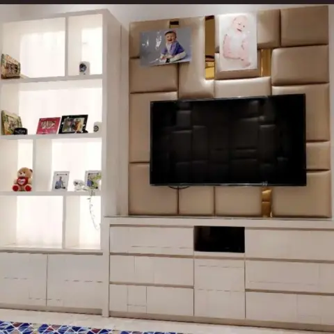 A living room TV wall with a diamond-tufted padded panel behind the screen, an open display shelf with books and a teddy bear, and a low storage console below