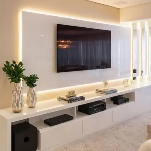 A living room TV wall with a glossy white backlit panel above a floating console styled with textured vases, stacked books, and a geometric-patterned rug.