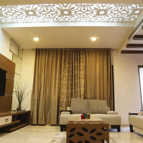 A living room with a backlit CNC-cut floral jaali ceiling panel above a wood-panelled TV wall, gold patterned curtains, and a carved wooden coffee table.