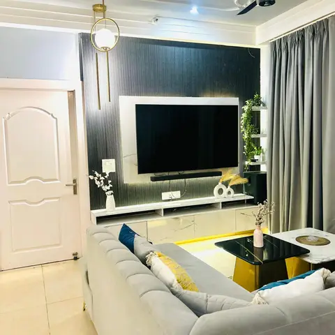A living room with a backlit fluted-panel TV wall, a gold ring pendant light, and a grey tufted sofa styled with blue and mustard cushions.