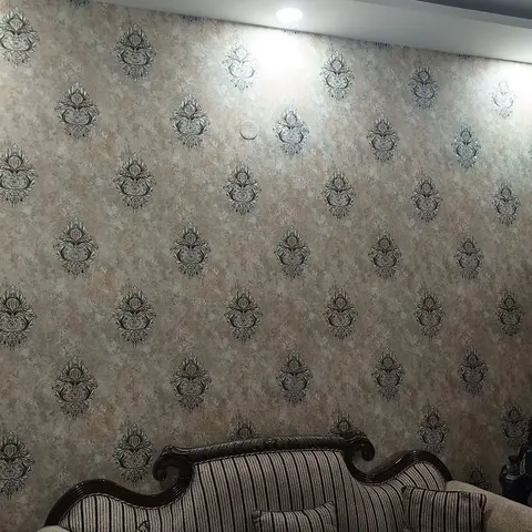 A living room with a bronze damask-print wallpaper feature wall behind a carved dark-wood sofa with striped upholstery