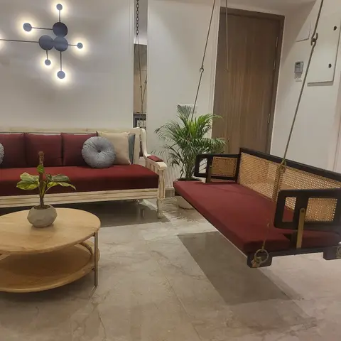 A living room with a cane-backed hanging jhula swing and a cream sofa with houndstooth pillows, both in matching maroon upholstery