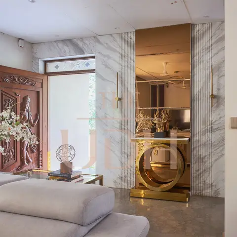 A living room with a carved wood entrance door bearing a deity relief, a marble-panelled wall behind a brass ring-base console table and bronze mirror
