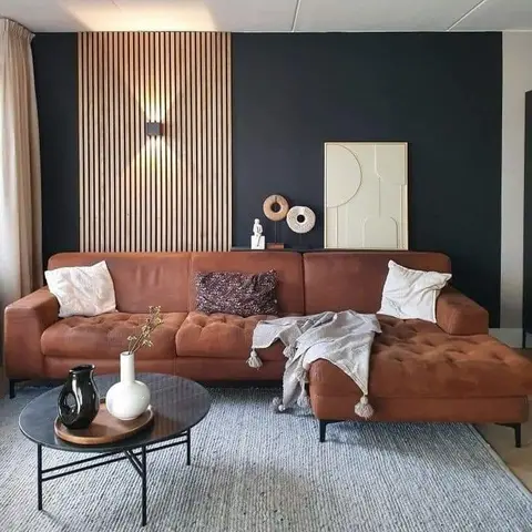 A living room with a cognac leather tufted sectional sofa, a round black coffee table, and a wood-slat accent wall with a sconce light against a navy backdrop.