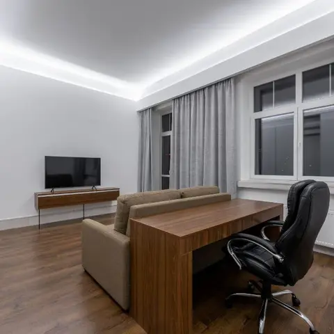 A living room with a cove-lit ceiling, floor-to-ceiling grey curtains, a wood TV console, and a taupe sofa paired with a wood desk and black office chair.