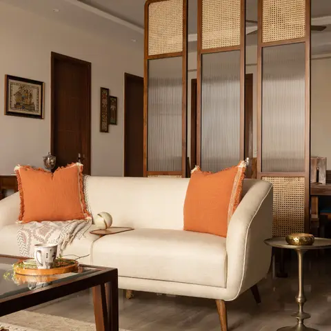 A living room with a cream mid-century sofa and orange linen cushions, in front of a cane-and-reeded-glass room-divider screen suspended from the ceiling