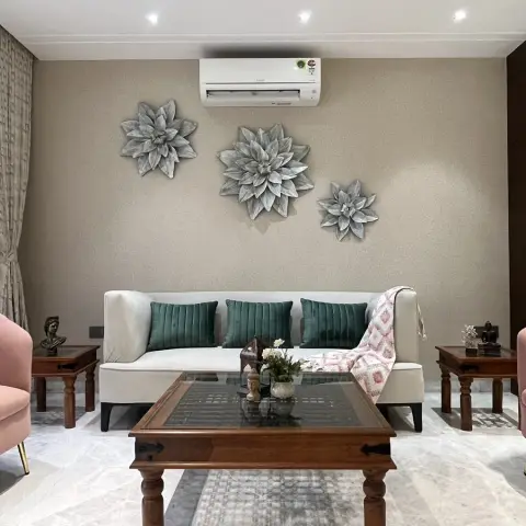 A living room with a cream sofa flanked by pink velvet armchairs, a carved wood coffee table, and a trio of oversized metal flower wall sculptures above.