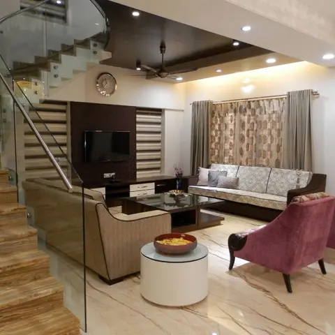 A living room with a curved glass-railed staircase, dark wood TV unit wall, beige sofa and maroon armchairs around a round glass-top coffee table on veined