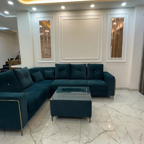 A living room with a deep-teal velvet L-shaped sofa with gold trim, a matching glass-top coffee table, and wall-panel moulding with mirror insets.