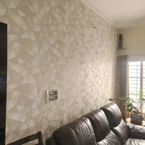 A living room with a faceted 3D geometric wallpaper feature wall, a dark brown leather sectional sofa, and sheer-and-blackout curtains by a grilled window.