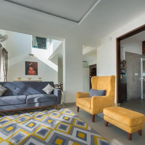 A living room with a grey tufted mid-century sofa and a mustard wingback armchair with ottoman on a yellow-and-grey geometric rug