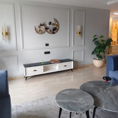 A living room with a grey moulded panel feature wall, brass sconces either side of a metal leaf wall sculpture, a white console with black marble top