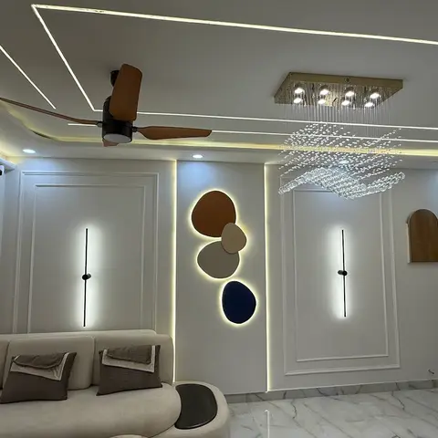 A living room with a linear-cove false ceiling, a crystal cluster chandelier, and a backlit pebble-shaped wall art panel between two panelled doors