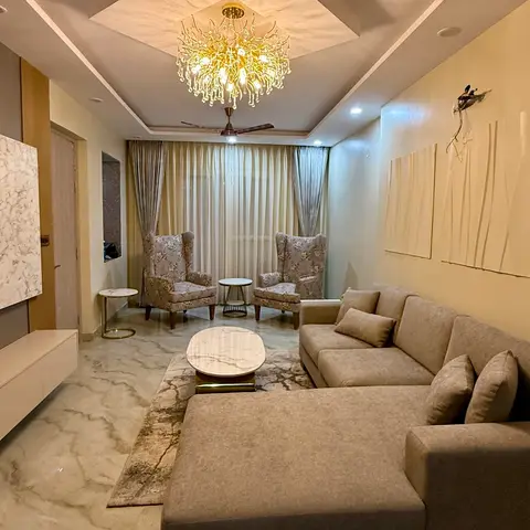 A living room with a marble-clad TV feature wall, a crystal chandelier in a cove-lit pop ceiling, floral wingback chairs by the curtained window
