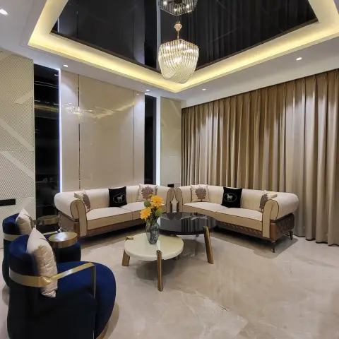 A living room with a mirror-finish pyramid false ceiling holding twin crystal chandeliers over a beige tufted sectional and navy-and-gold accent armchairs on